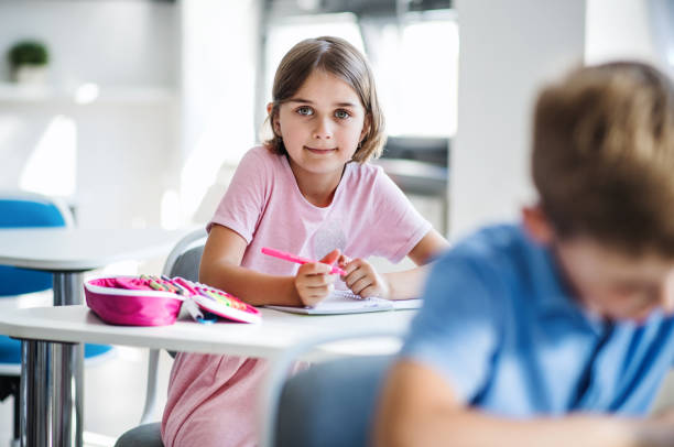 une petite fille d'école s'asseyant au bureau dans la salle de classe, regardant l'appareil-photo. - schoolgirl little girls crayon human face photos et images de collection
