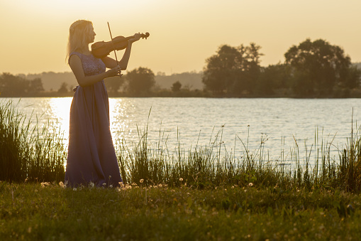 romantic silhouette of young woman with a violin at dawn on river bank, elegant girl playing a musical instrument on nature, concept music and inspiration