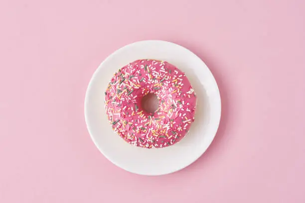 donat decorated sprinkles and icing in white plate on pink background. Creative and minimalis food concept, top view flat lay