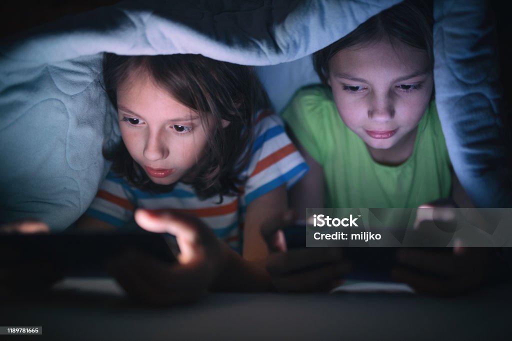 Boy and girl playing games on mobile phone in their bed Boy and girl playing games on mobile phones while lying on bed in bedroom under the blanket. They are spending some nice time together that makes them happy. Child Stock Photo