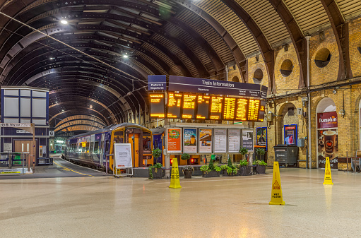 York Railway Station at night.  An electronic departure board dominates the scene as a train waits by the platform. A curved canopy is overhead.
