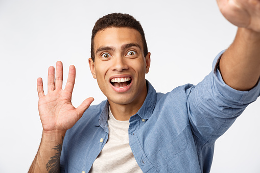 Surprised and impressed man didnt expact see someone as holding camera, waving hand in hello or hi gesture, pop eyes fascinated, open mouth and smiling fascinated, white background astonished.