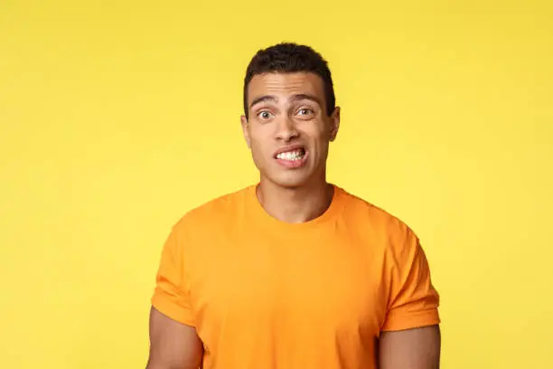 Photo of Yikes, awkward. Embarrassed young handsome man caught on lie, feel indesicive and slightly worried, cringe, smiling nervously and look camera with reluctance, stand yellow background
