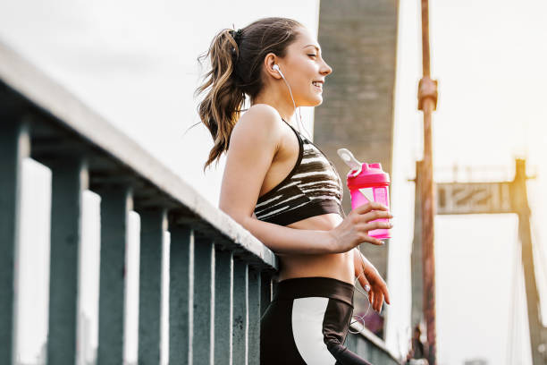 Happy fitness woman drinking water and smiling outdoors Portrait of happy attractive fitness girl in stylish sportswear drinking water from shaker while standing on bridge. Beautiful slender sports woman listening to music and smiling. Workout outdoors cocktail shaker stock pictures, royalty-free photos & images