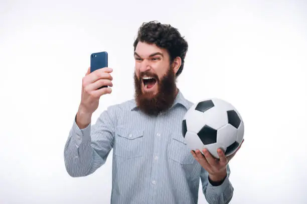 Screaming bearded man in casual looking at smartphone and holding soccerball