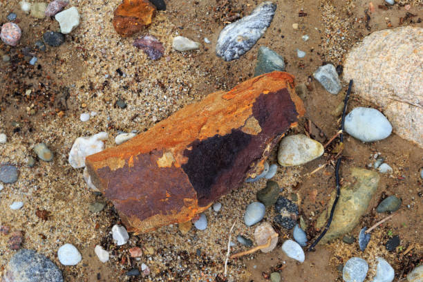 Large stone of rust colored, surrounded by smaller stones on the seashore.