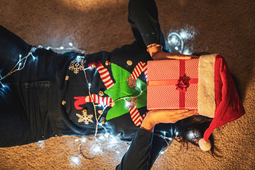 Woman laying on back over the carpet with Christmas deocation, she holding Christmas present in front of her face.