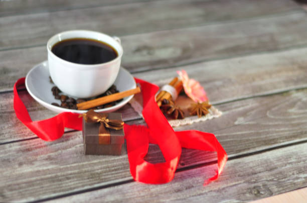 istock Ceramic cup on a saucer with black coffee, decorated with grains, anise, a flower bud and cinnamon, next to a gift box and a scarlet ribbon around the composition. 1189882632