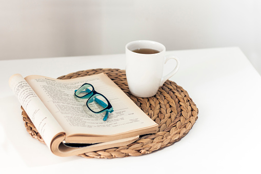 open book and glasses, lemon tea in a white ceramic cup on a wicker stand on the table, cozy home interior