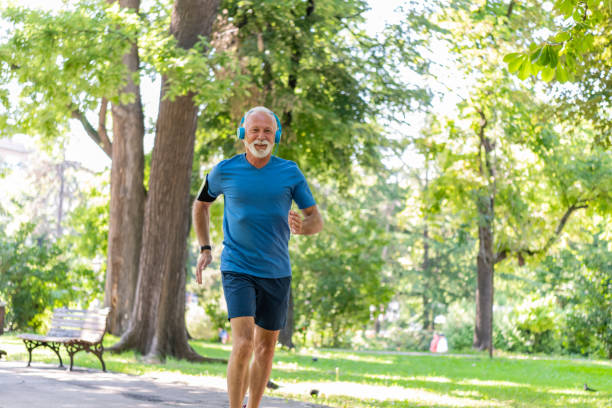 Active Senior Athlete Man is Running in the Public Park. Older Sportsman with Grey Hair and Beard with Wearable Computer is Jogging in the Park and Listening to Music.
