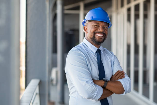 Successful contractor at building site Portrait of african american man architect at building site with folded arms looking at camera. Confident construction manager in formal clothing wearing blue hardhat. Successful mature civil engineer at construction site with copy space. civil engineer stock pictures, royalty-free photos & images