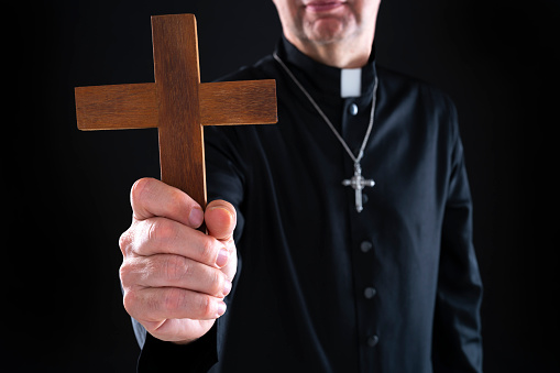 Priest bald with cassock holding crucifix wooden cross and bible on black background