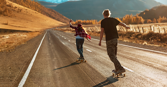 Young couple is riding on longboards by straight mountain road at sunset light