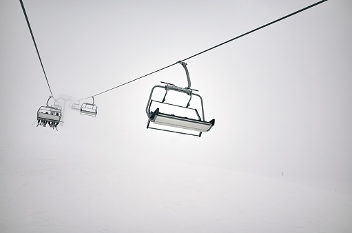 Skiers on the chair lift ropeway winter resort, clouds and mist, gray cloudy sky and slope background, silhouette