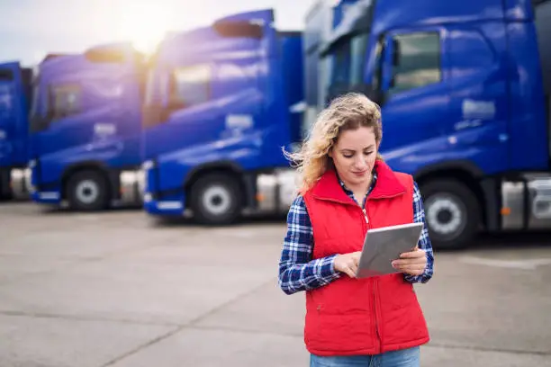 Truck driver holding tablet and checking route for new destination. In background parked truck vehicles. Transportation service.