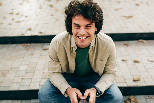 Portrait of smiling young man standing outdoors, typing messages on mobile phone. Young male with curly hair sittiing on the stairs in the city street resting outside, browsing on his smart phone