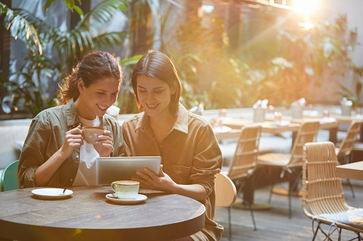 Portrait of two contemporary women looking at tablet screen while enjoying meeting in sunlit care terrace, copy space