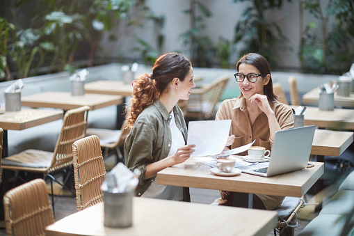 Portrait of two young businesswomen discussing project while sitting at table in designer cafe, copy space