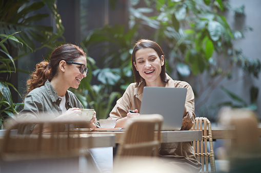 Portrait of two young women laughing happily while using laptop on outdoor cafe terrace decorated with plants, copy space