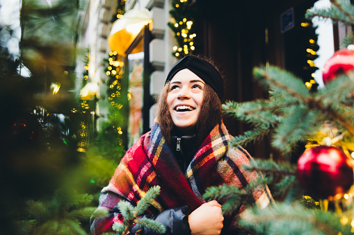 Young beautiful woman with curly hair and a big warm scarf walking in the city, enjoying the Christmas decorations everywhere and choosing the perfect Christmas Tree for her home