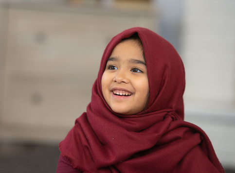 A sweet young Muslim girl sits in the comfort of her home while posing for a portrait.  She is dressed casually and wearing a Hijab.  She is looking up to the left and smiling.