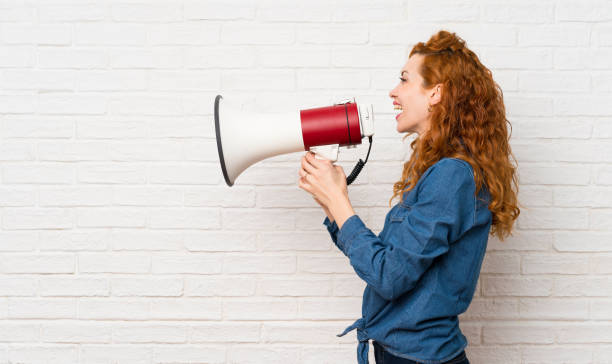 istock Redhead woman over white brick wall shouting through a megaphone 1189996440