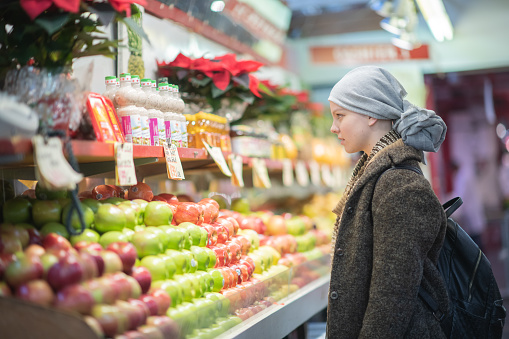 A young Caucasian woman with Cancer and dressed casually in a wool coat and a head scarf, walks the aisles of the grocery store shopping.  She is down the produce aisle and looking at fruit.