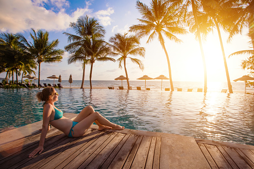Happy relaxing woman sitting near swimming pool and looking on sunset sea and coconut palm trees. Luxury resort hotel with deck chairs. Vacation travel to tropical exotic island country.