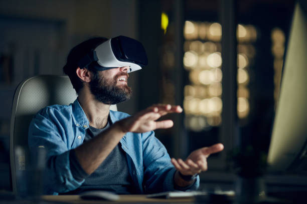 Excited young bearded man sitting in front of computer and gesturing hands while testing new app via VR device Man testing new app via VR device virtual reality simulator stock pictures, royalty-free photos & images