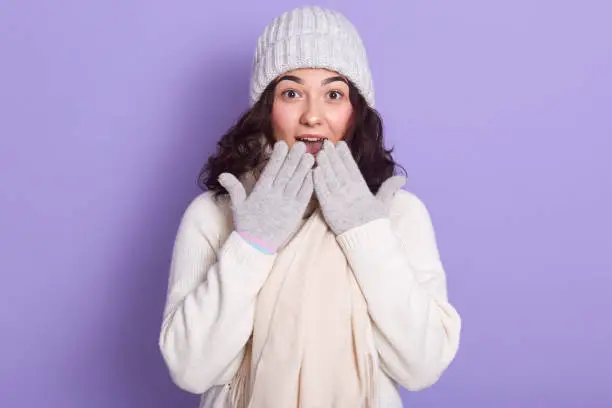 Photo of Young attractive girl wearing white sweater, warm cap, scarf and gloves, having light makeup, covering her mouth with her hands, looking with big eyes in surprise, isolated over lilac background.