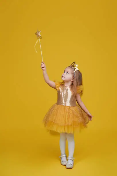 Joyful little girl with long hair in a tulle golden dress and princess crown holding a magic wand  on yellow background. Celebrating a colorful carnival for kids, expressing positive birthday