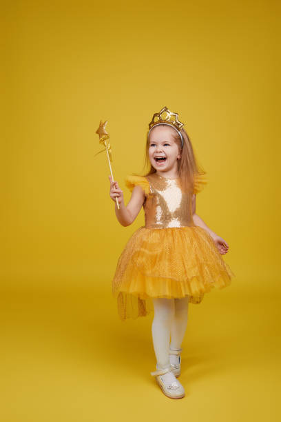 Little princess in golden dress Joyful little girl with long hair in a tulle golden dress and princess crown holding a magic wand  on yellow background. Celebrating a colorful carnival for kids, expressing positive birthday carnival children stock pictures, royalty-free photos & images