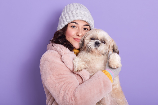 Dark haired woman wearing faux fur coat andwarm white cap, hugging her cute puppy isolated over lilac background, looking directly at camera, attractive lady spending free time with her favourite pet.