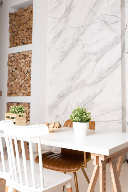 Bright dining room with big white wooden table. Marble wall on the background, firewood for a fireplace