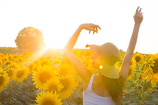 A happy, beautiful young girl in a straw hat is standing in a large field of sunflowers. Summer time. Back view. The beautiful girl in Sunflowers field , emotional girl, enjoying nature and laughing on summer sunflower field. Sunflare, sunbeams, glow sun. Backlit, hiding behind flowers