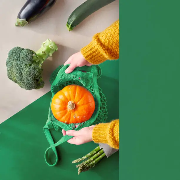 Eco friendly zero waste flat lay with hands holding broccoli and string bag with vibrant orange pumpkin. Fall flat lay with vegetables on two color paper background, craft paper and green.