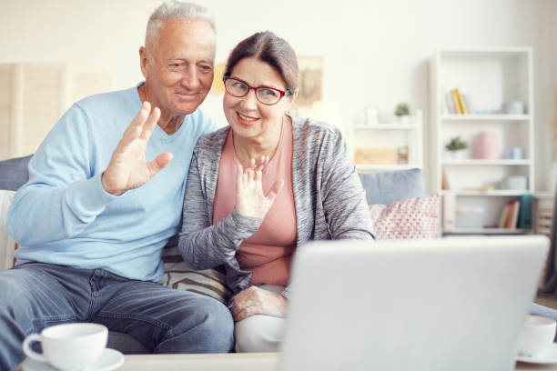 istock Cheerful modern grandparents sitting on sofa and waving hands to laptop webcam while communicating online 1190023545