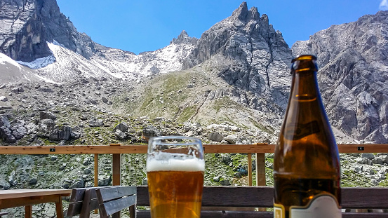 Bottle of beer and a pint glass standing on a wooden table in a mountain cottage. Tall Lienz Dolomites are the backdrop. Sharp peaks look dangerous.  Relaxation after hike. Enjoying the view and drink