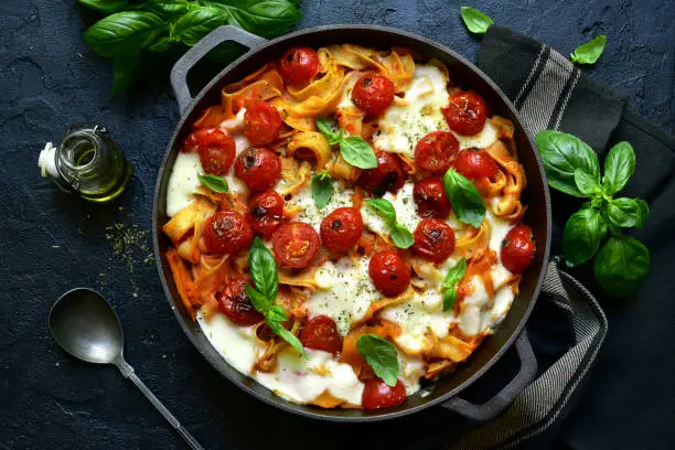 Pasta casserole with tomatoes and mozzarella cheese in a cast iron pan on a dark slate, stone or concrete background. Top view with copy space.