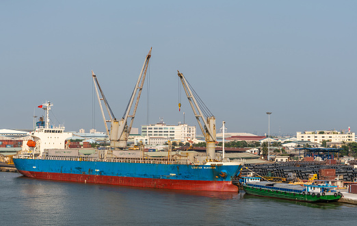 Ho Chi Minh City, Vietnam - March 13, 2019: Tan Thuan port on Song Sai Gon river at sunset. Blue-red Lizstar Success container vessel at quay where rolls of black metal wire are stored. Small barge in front.