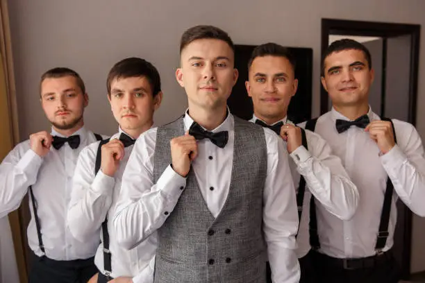 Elegant groom and groomsmen with suspender holding bow tie at wedding day. Group portrait of groom and his friends