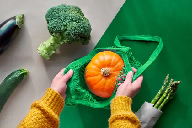 Eco friendly zero waste flat lay with hands holding broccoli and string bag with orange pumpkin.Autumn or Spring top view from above with vegetables on two color paper background, craft and green.