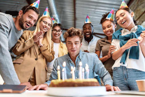 Corporate party. Young happy man blowing candles on cake while celebrating birthday with cheerful colleagues wearing party hats. Birthday concept. Corporate party