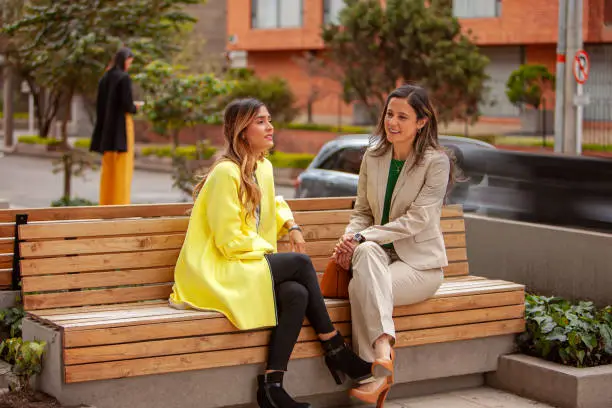 A couple of Latin American lady colleagues are seen seated outside their office building enjoying the morning sunshine. They are chatting with each other. One of them is more formally attired in a suit; while the other is more casually dressed. Their hair is untied and has been styled with highlights. Horizontal format; Copy space.