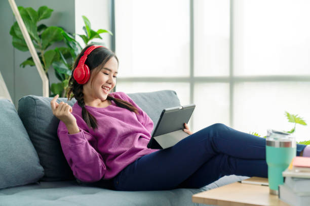 candid of asian woman using tablet for watching online movie stream mobile device on sofa at living room. young happy girl enjoying with entertainment media online on weekend. - digital tablet women enjoyment happiness imagens e fotografias de stock