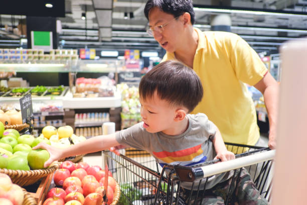 Asian father and son are shopping in supermarket. Cute little 3 years old toddler boy child sit in shopping cart choosing fruits (green apple) in grocery store stock photo