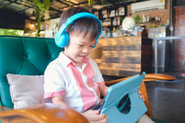 asian 3 - 4 years old toddler boy child smiling while sitting in armchair using tablet pc computer, gadget addicted children, learning tablet for kids - toddler music asian ethnicity child imagens e fotografias de stock