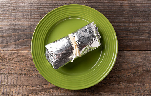 Burrito wrapped in foil and neatly tied closed with a corn husk served on a green plate on a reclaimed wood table.