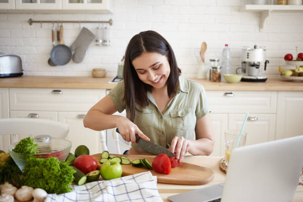 istock Charming brunette girl in her twenties learning how to make vegan lasagne, using sharp knife while cutting bell pepper on cooking board, siting in front of laptop, surrounded with all ingredients 1190100589