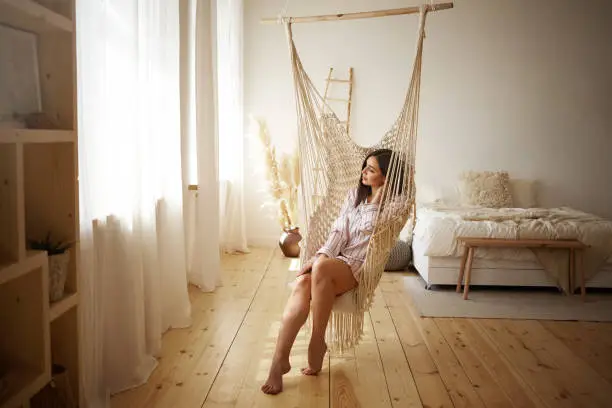 Photo of Beauty, style, rest and relaxation concept. Picture of fashionable young plus size female enjoying slow lazy weekend morning at home, sitting barefooted in hanging chair in light spacious bedroom